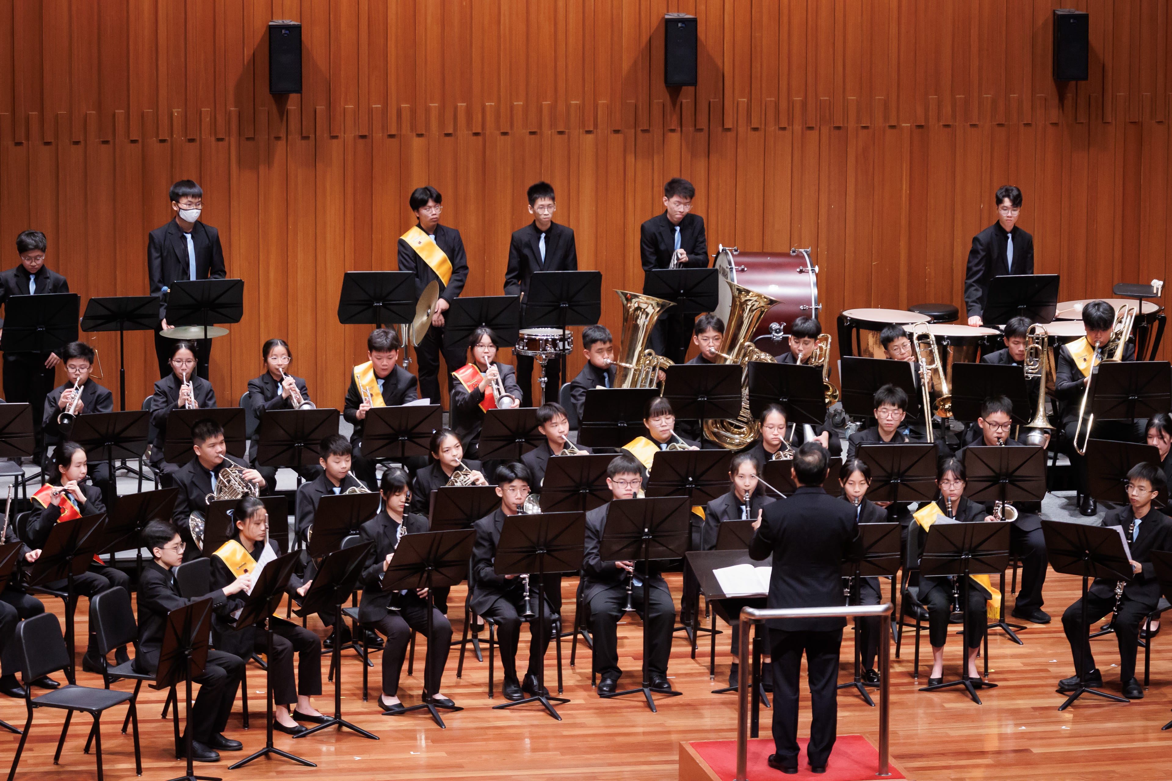 A youth wind band in black uniforms performs on a wooden stage, led by a conductor standing at a podium.